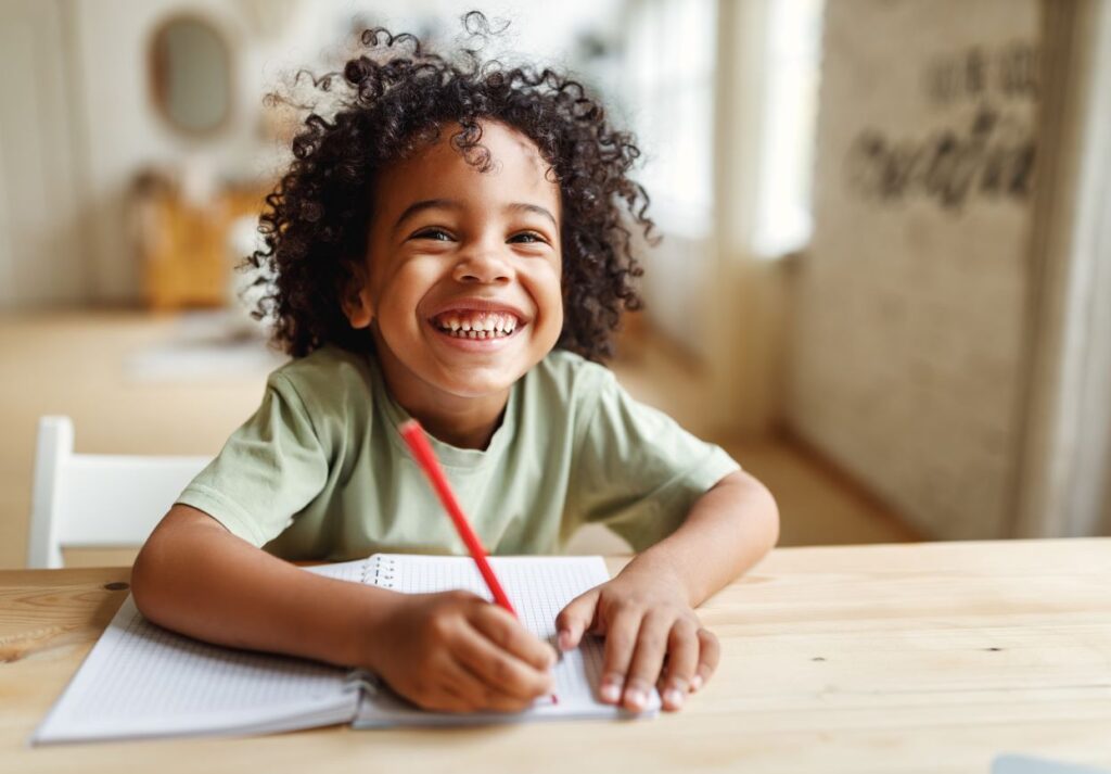 A little boy at school smiling and holding a red pencil.